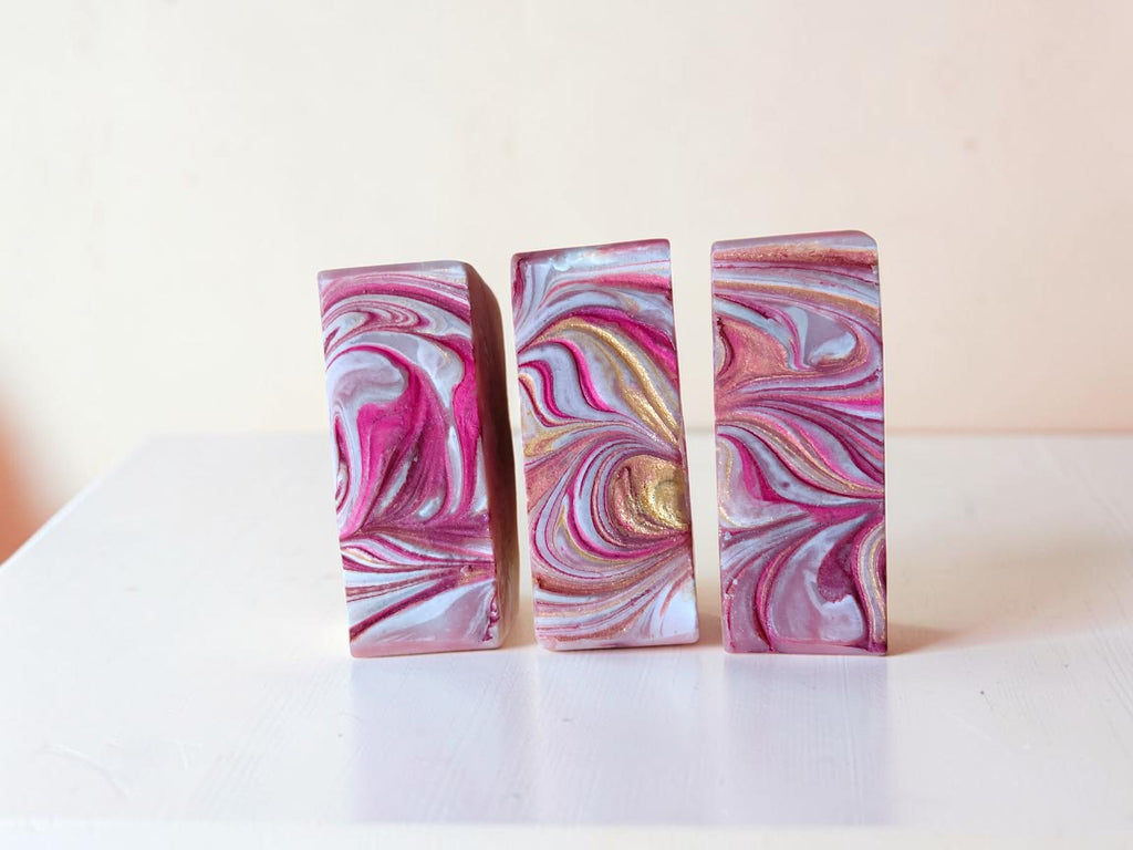 Three rectangular bars of soap with a pink, white, and gold marbled swirl pattern, standing upright on a white surface against a light beige background.