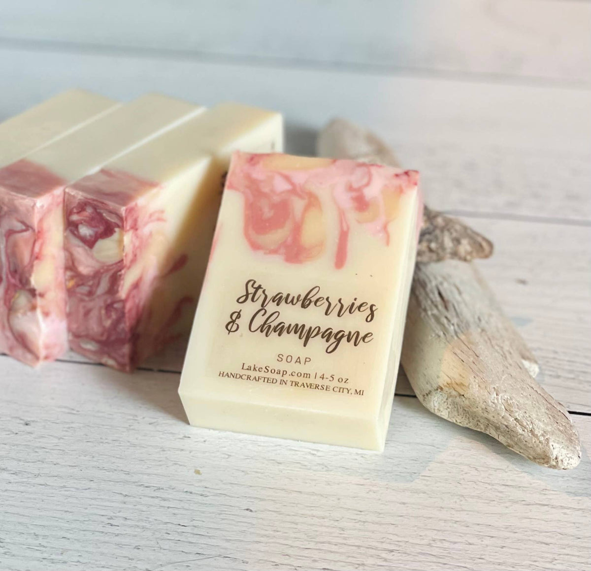 A handmade bar of soap labeled 'Strawberries & Champagne' stands upright on a whitewashed wooden surface, with two additional marbled soap bars and driftwood props in the background.