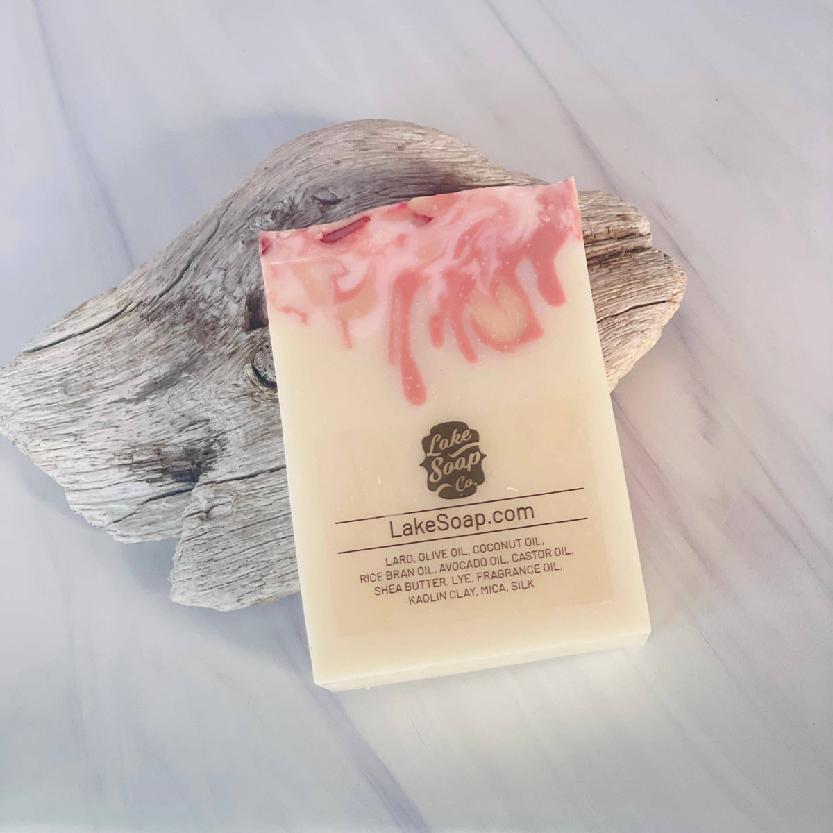 A rectangular bar of Lake Soap handmade soap with pink and cream swirls, resting on a piece of weathered driftwood over a light marble surface.